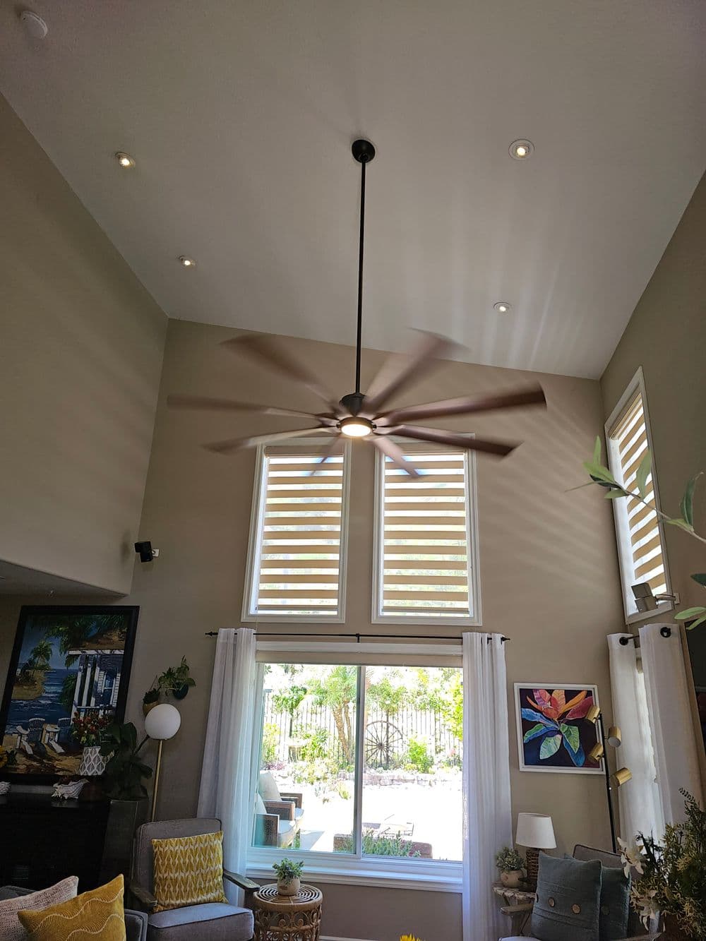 Modern living room with ceiling fan, natural light, and large windows overlooking greenery.