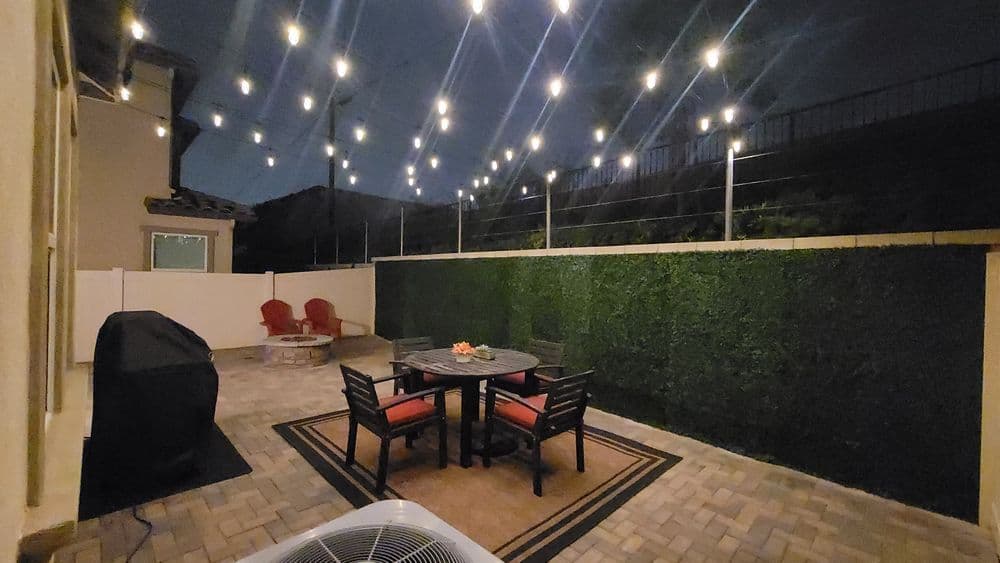 Outdoor patio with string lights, dining table, and seating area surrounded by greenery at night.